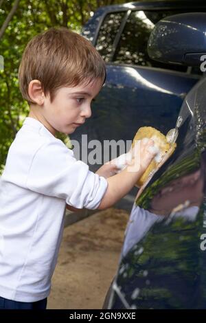un jeune garçon lavant une voiture avec une éponge humide pour une idée d'activité estivale. Banque D'Images