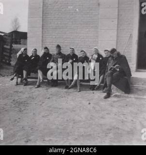 Rare moment historique de la Seconde Guerre mondiale. Deux soldats sont assis parmi des infirmières sur le banc devant une maison ou une église. Les infirmières sourient, mangent, se reposent. Un soldat se concentre pour le photographe, l'autre ne donne pas un lancer qu'il lit. Source: Photographie originale (sujet militaire) Banque D'Images