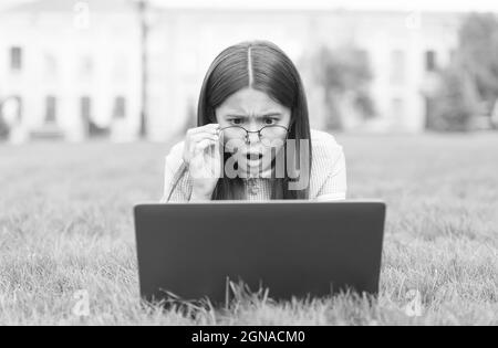Oh non. Retour à l'école. Jeune fille utiliser l'ordinateur sur l'herbe verte dans le parc. Enfant dans les lunettes avec le carnet. Nouvelle technologie dans la vie moderne. Étude. Surpris Banque D'Images