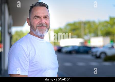 Homme sportif expérimenté et confiant avec une barbe debout à l'extérieur dans une rue tranquille tournant pour regarder attentivement la caméra avec un espace de travail Banque D'Images