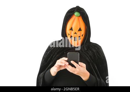 Personne vêtue de masque citrouille Jack-O.-lanterne et cape en velours à capuchon, regardant son téléphone mobile, sur fond blanc. Carnaval, Halloween et jour o Banque D'Images