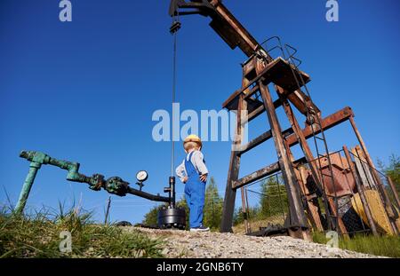 Adorable enfant dans une combinaison de travail à proximité d'un cric à pompe à pétrole sous un beau ciel bleu. Petit garçon en construction casque de surveillance du travail de la pompe à huile de rocker-machine dans le champ de pétrole. Banque D'Images