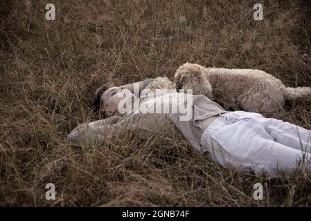 Portrait d'un jeune homme de 18 ans, allongé avec un chien, doux-enduit Wheaten Terrier, dans l'herbe sèche, dans les rayons du soleil couchant. Banque D'Images