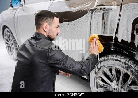 Un jeune homme lave-auto à l'extérieur. Beau conducteur de nettoyage automobile, à l'aide d'éponge et de savon. Banque D'Images