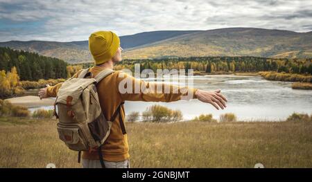 Touriste dans un chapeau jaune et chandail avec un sac à dos est debout avec ses bras étendus sur les côtés en face d'une rivière, la forêt dorée d'automne Banque D'Images
