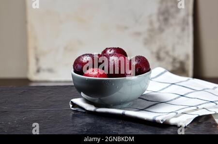 Prunes rouges mûres dans un bol avec gouttes d'eau. Fruits juteux sur fond en bois, gros plan. Des aliments sains sur une table de cuisine sombre Banque D'Images