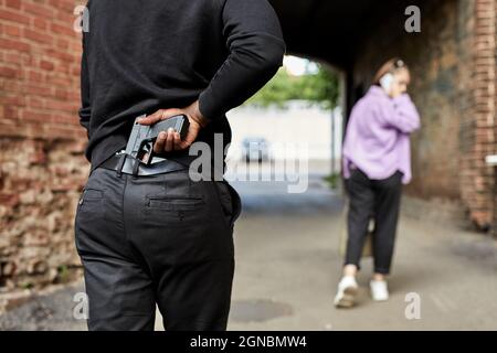 Vue arrière sur un homme méconnaissable transportant un fusil et suivant une jeune femme marchant dans la rue seule. Violence contre les femmes, le concept de viol. De vraies personnes. V Banque D'Images