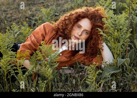 redhead femme couchée sur l'herbe entourée de fantastiques plantes de fées, femme maurille avec des cheveux rouges naturels posant à l'appareil photo, portant une chemise orange décontractée, dans Banque D'Images