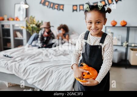 Portrait de bonne fille afro-américaine tenant la citrouille sculptée et souriant à l'appareil photo tout en se tenant debout dans la pièce avec des décorations d'Halloween, un espace pour les copies Banque D'Images