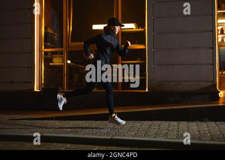 vue latérale sur la jeune athlète caucasienne, femme qui court devant un café de la ville la nuit, sport en plein air. une femme motivée et concentrée est en avance, leader actif Banque D'Images