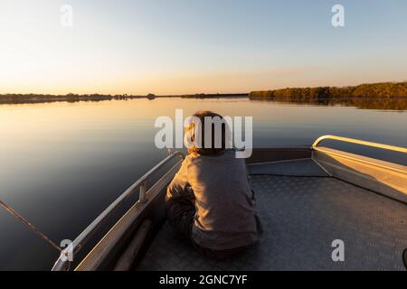 Jeune garçon assis à la poupe d'un bateau sur le delta d'Okavango au coucher du soleil Banque D'Images