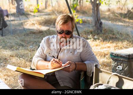 Homme barbu portant des lunettes de soleil assis sur une chaise dans un camp de safari, écrivant un journal Banque D'Images