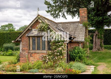 Très ancienne petite maison de jardin en brique rouge avec toit en ardoise, fenêtres à meneaux et rose grimpant. Banque D'Images