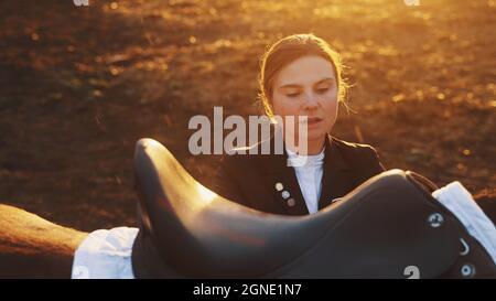 l'accent est mis sur une jeune femme élégante avec un beau visage qui se tient à côté d'un cheval et met en place une selle sur un sol inégal. Photo de haute qualité Banque D'Images