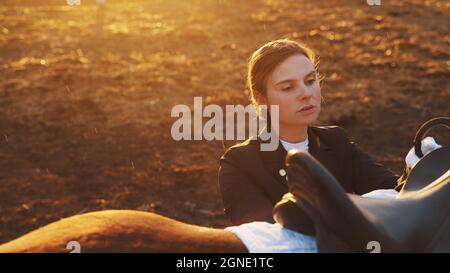 le foyer sur une jeune femme élégante avec un beau visage se tient à côté d'un cheval et met en place une selle. Photo de haute qualité Banque D'Images