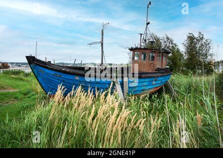 Vieux bateau de pêche garé sur un pré. En arrière-plan un petit port sur la côte Baltique Banque D'Images