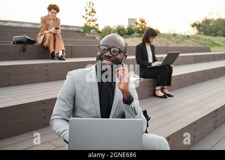 Portrait horizontal moyen d'un costume d'homme d'affaires afro-américain à barbe en toute confiance assis sur un banc à l'extérieur avec un ordinateur portable parlant au téléphone avec un co-worke Banque D'Images