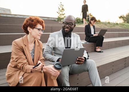 Portrait horizontal d'un homme afro-américain moderne assis à l'extérieur avec son collègue qui lui montre quelque chose sur son ordinateur portable Banque D'Images
