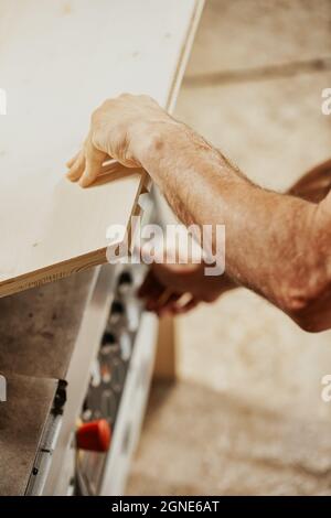 Carpenter soulevant une feuille de bois pour accéder aux commandes d'une machine à bois ci-dessous dans un atelier de menuiserie en gros plan sur ses mains Banque D'Images