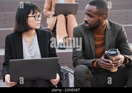 Portrait horizontal moyen tourné de jeunes Afro-américains et asiatiques portant des vêtements élégants assis à l'extérieur travaillant ensemble sur les relations publiques d'affaires Banque D'Images