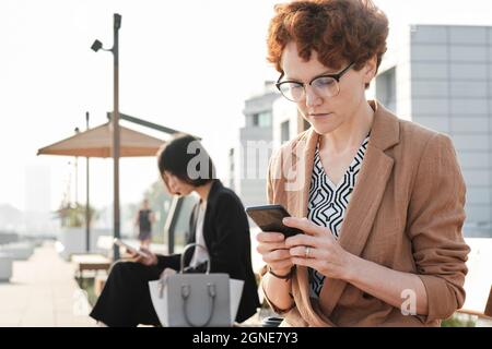 Portrait horizontal moyen de femme mûre avec de courts cheveux rouges assis sur un banc à l'extérieur surfer sur Internet sur un smartphone Banque D'Images
