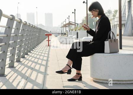 Vue latérale horizontale d'une jeune femme asiatique attrayante portant une tenue élégante et à la mode, assise à l'extérieur surf sur Internet sur un smartphone, Copy spa Banque D'Images