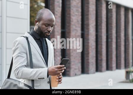 Portrait horizontal moyen d'un employé de bureau américain moderne d'Afrique portant des écouteurs marchant à l'extérieur en choisissant une liste de lecture sur son smartphone Banque D'Images