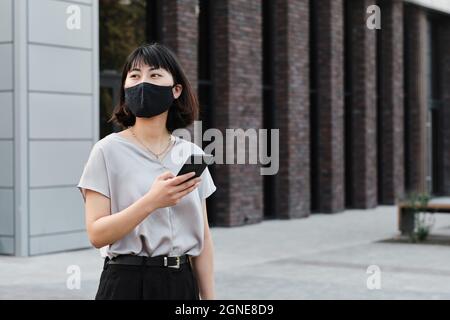 Portrait horizontal moyen d'une jeune femme asiatique portant un masque de protection sur le visage tenant un smartphone debout dans le quartier des affaires et regardant loin Banque D'Images