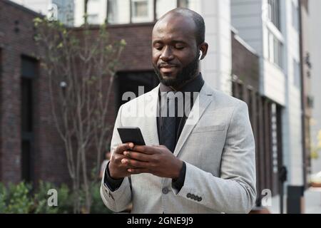 Portrait horizontal moyen d'un homme afro-américain joyeux avec une barbe sur le visage portant des écouteurs debout à l'extérieur en choisissant une liste de lecture pour écouter sur ses s Banque D'Images