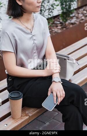 Vue en grand angle verticale d'une femme asiatique élégante et méconnaissable assise sur un banc tenant son smartphone pendant une pause café Banque D'Images