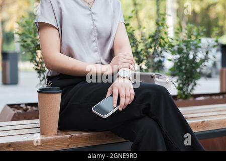 Photo horizontale en coupe moyenne d'une femme élégante et méconnaissable assise sur un banc tenant un smartphone pendant une pause café Banque D'Images