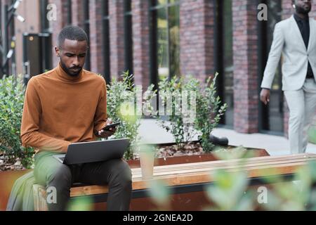 Portrait horizontal d'un jeune travailleur de bureau afro-américain assis à l'extérieur, concentré sur les tâches de travail à l'aide d'un ordinateur portable Banque D'Images