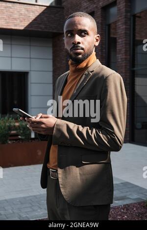 Portrait vertical moyen d'un jeune Africain américain avec barbe sur le visage portant un costume et un pull à col roulé debout à l'extérieur tenant un smartphone Loo Banque D'Images