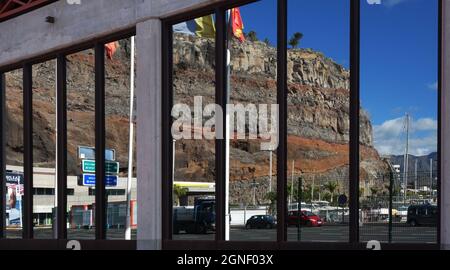 La réflexion sur une fenêtre de magasin, où nous voyons le port, les montagnes, le ciel, les bateaux, les drapeaux,Voitures, dans le port de San Sebastian, îles Canaries Banque D'Images