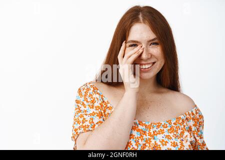 Gros plan de la belle fille heureuse avec les cheveux rouges et la peau naturelle du visage, riant, touchant son visage avec une main et souriant coquettish à l'appareil photo Banque D'Images