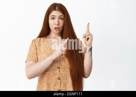 Surprise redhead fille poitns dans le coin supérieur droit, pose la question, se tient contre fond blanc Banque D'Images