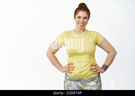 Souriant, le curvy fille s'entraîner dans des vêtements de fitness, tenant les mains à la taille et regardant motivé, se tenant sur fond blanc Banque D'Images