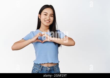 Sourire brune asiatique fille montre coeur signe, kokoro geste, debout sur fond blanc Banque D'Images