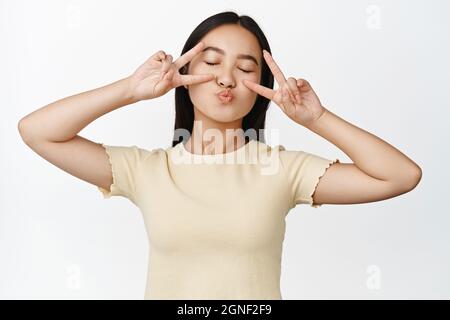 Jolie fille asiatique rêveuse, yeux proches et lèvres de mouvement, faisant des baisers visage avec des signes de paix, debout dans un t-shirt jaune sur fond blanc Banque D'Images