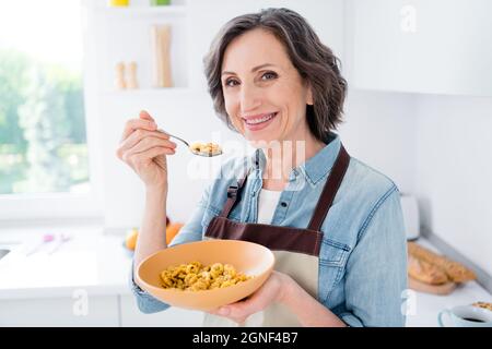 Photo de gaie positive joyeuse vieille femme tenir main assiette cuillère céréales à l'intérieur maison cuisine Banque D'Images