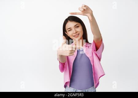 Une jeune fille créative souriante à la recherche d'un angle parfait, prenant des photos, en se faisant un clin d'œil et regardant à travers le cadre de la main, se tenant sur un fond blanc Banque D'Images