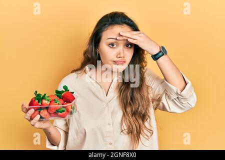 Jeune fille hispanique tenant des fraises inquiètes et stressées au sujet d'un problème de main sur le front, nerveux et anxieux pour la crise Banque D'Images