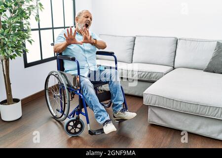 Beau homme senior assis sur un fauteuil roulant dans le salon peur et terrifié avec l'expression de peur stop geste avec les mains, criant dans le choc. PAN Banque D'Images