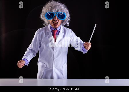 Professeur de science en colère en manteau blanc avec des cheveux unkempt dans des lunettes drôles d'oeil tenant une baguette pour pointer au tableau noir Banque D'Images