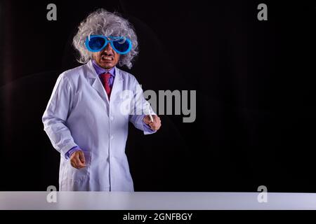 Professeur de science en colère en manteau blanc avec des cheveux unkempt dans des lunettes drôles d'oeil tenant une baguette pour pointer au tableau noir Banque D'Images