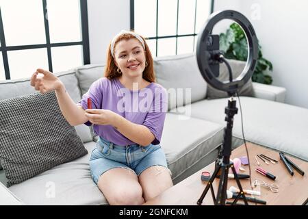 Jeune femme à tête rouge faisant le maquillage tutorial en utilisant le smartphone à la maison Banque D'Images