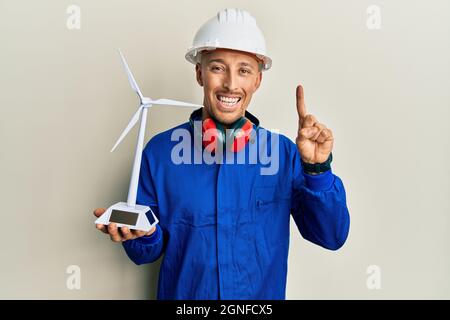 Homme d'ingénieur chauve avec barbe tenant un moulin solaire pour l'électricité renouvelable souriant avec une idée ou une question pointant du doigt avec visage heureux, nombre Banque D'Images