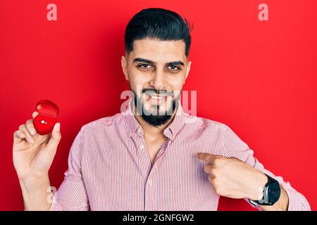 Beau homme avec la barbe tenant l'anneau d'engagement pour la proposition pointant du doigt à un soi souriant heureux et fier Banque D'Images