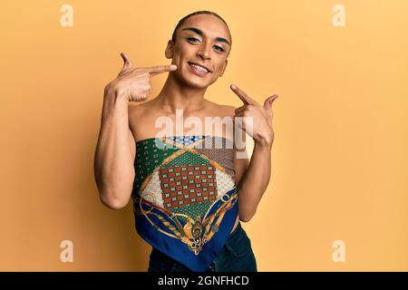 Hispanique homme portant le maquillage et les cheveux longs portant des femmes haut sourire gai montrant et pointant avec les doigts dents et la bouche. Santé dentaire concep Banque D'Images
