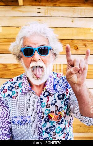 Homme senior faisant une expression drôle et faisant des gestes contre le mur en bois. Portrait de l'homme âgé en chemise et lunettes de soleil funky. Vieux gesticulations avec ha Banque D'Images